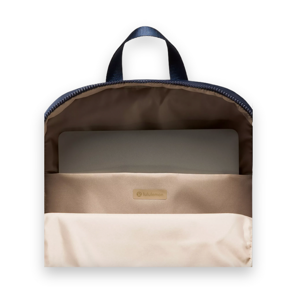 Open view of a Lululemon backpack showing the beige interior lining, a zippered compartment, a laptop inside, and a navy blue top handle against a light gray background.