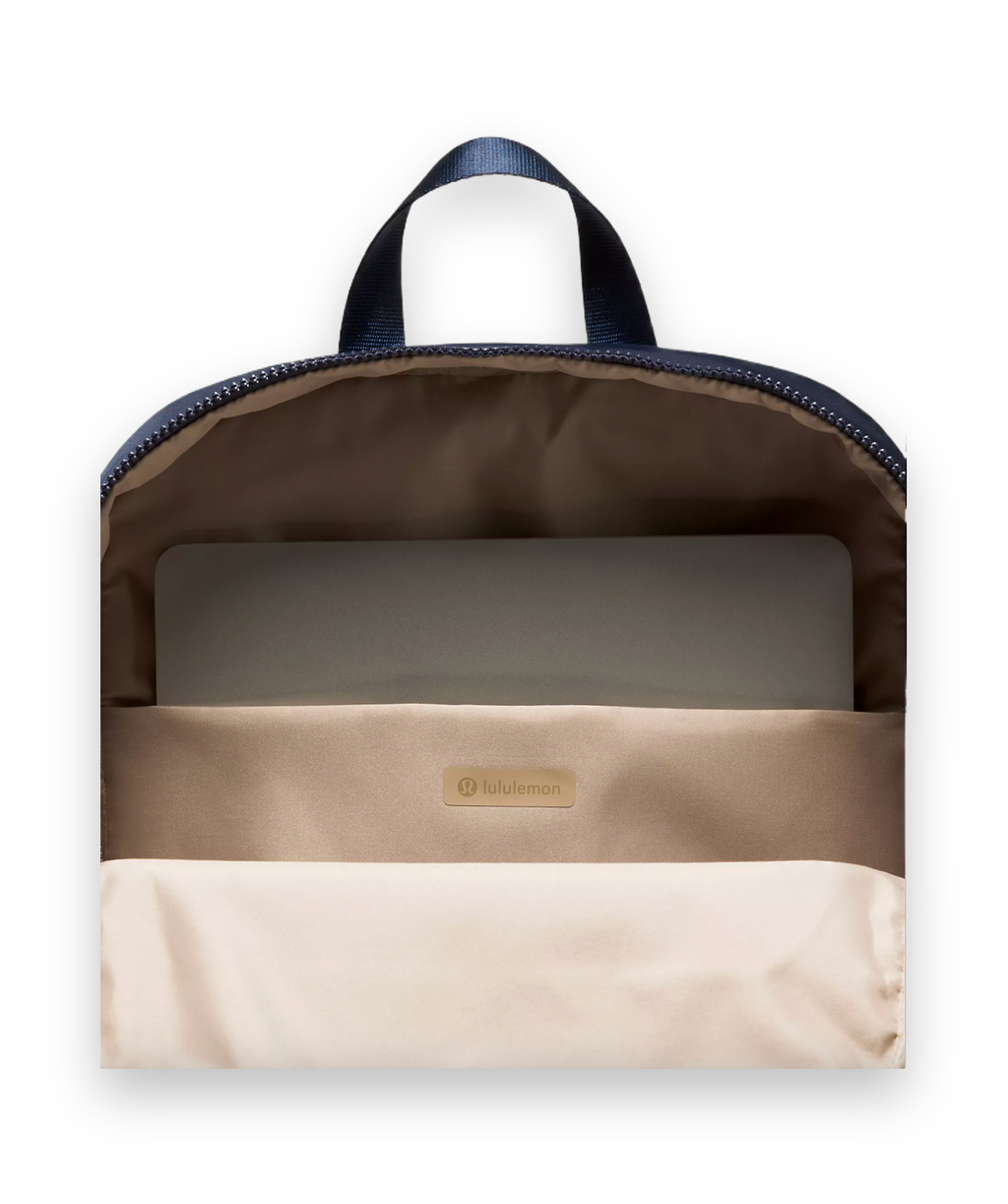 Open view of a Lululemon backpack showing the beige interior lining, a zippered compartment, a laptop inside, and a navy blue top handle against a light gray background.